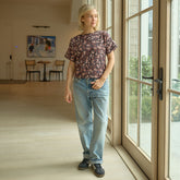 Woman wearing a brown floral top with blue jeans, standing by a large window in a room with wooden flooring.