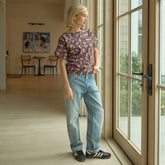 Woman standing by a window wearing a floral top and jeans, looking out.