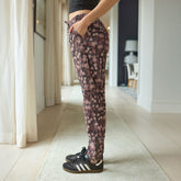 Side view of woman wearing brown floral women's joggers with gold zipper details standing in a hallway with her hand in her pocket. 