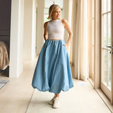 Woman wearing a white tank top and light blue bubble skirt standing in a bright room with large windows.