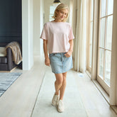 Woman wearing a boxy, baby pink t-shirt and denim skirt standing in a modern living room.
