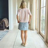 Back view of woman wearing a pink shirt and denim skirt standing in a room with large windows.
