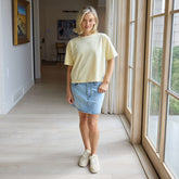 Woman wearing Albion's Perfect Tee—a yellow t-shirt with denim skirt standing in a room with large windows.