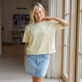 Woman wearing a casual butter yellow t-shirt and denim skirt standing in a room with large windows.