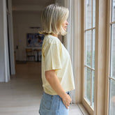 Woman facing the side in The Perfect Tee in yellow with denim skirt standing by a window in a home.