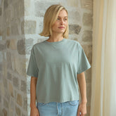 Woman wearing a light green, boxy t-shirt and denim skirt standing against a stone wall.