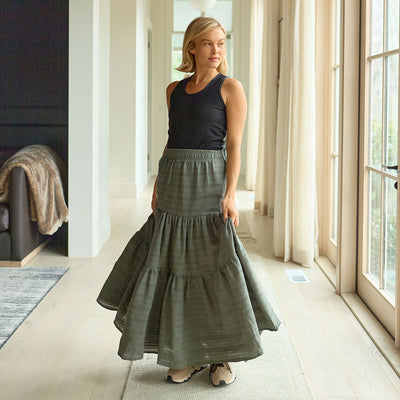 Woman wearing a long green skirt and black top in a bright room with large windows.