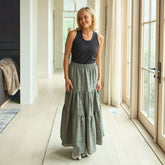 Woman wearing a black sleeveless top and green tiered maxi skirt standing in a bright room with large windows.