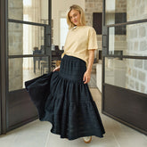 Woman wearing a black tiered maxi skirt and beige top in a modern indoor setting.