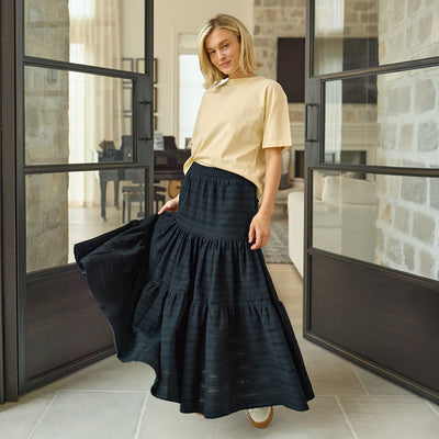 Woman wearing a black tiered maxi skirt and beige top in a modern indoor setting.
