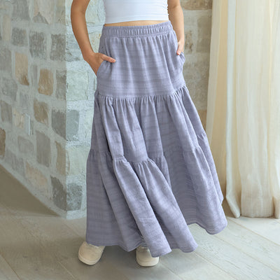 Woman wearing a long pebble blue maxi skirt with a stone wall and curtain in the background