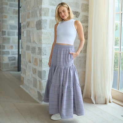 Woman wearing a white sleeveless top and periwinkle tiered maxi skirt standing in a room with stone walls and large windows.