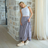 Woman wearing a white top and light blue maxi skirt standing against a stone wall.