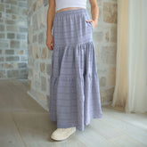 Woman wearing a long, tiered periwinkle maxi skirt with a stone wall background
