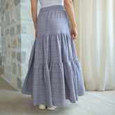 Woman wearing a long, light blue skirt with a stone wall and white curtain in the background.