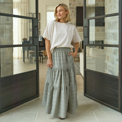 Woman in a white t-shirt and taupe maxi skirt standing in a modern interior setting with large glass doors.