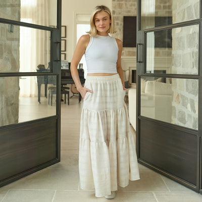 Woman in a white top and off white maxi skirt standing in a modern interior setting.