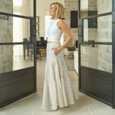 Woman in a white maxi skirt standing in a modern interior setting with large glass doors.