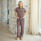 Woman wearing a brown floral jumpsuit standing in a room with stone walls and curtains.