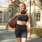 A woman stands on an outdoor basketball court wearing a matching black activewear set consisting of a sports bra and high-waisted shorts, holding a basketball and wearing black headphones around her neck.