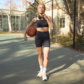 A woman stands on an outdoor basketball court wearing a black sports bra and matching high-waisted biker shorts, holding a basketball and headphones. The background shows a chain-link fence, trees, and buildings in soft focus.