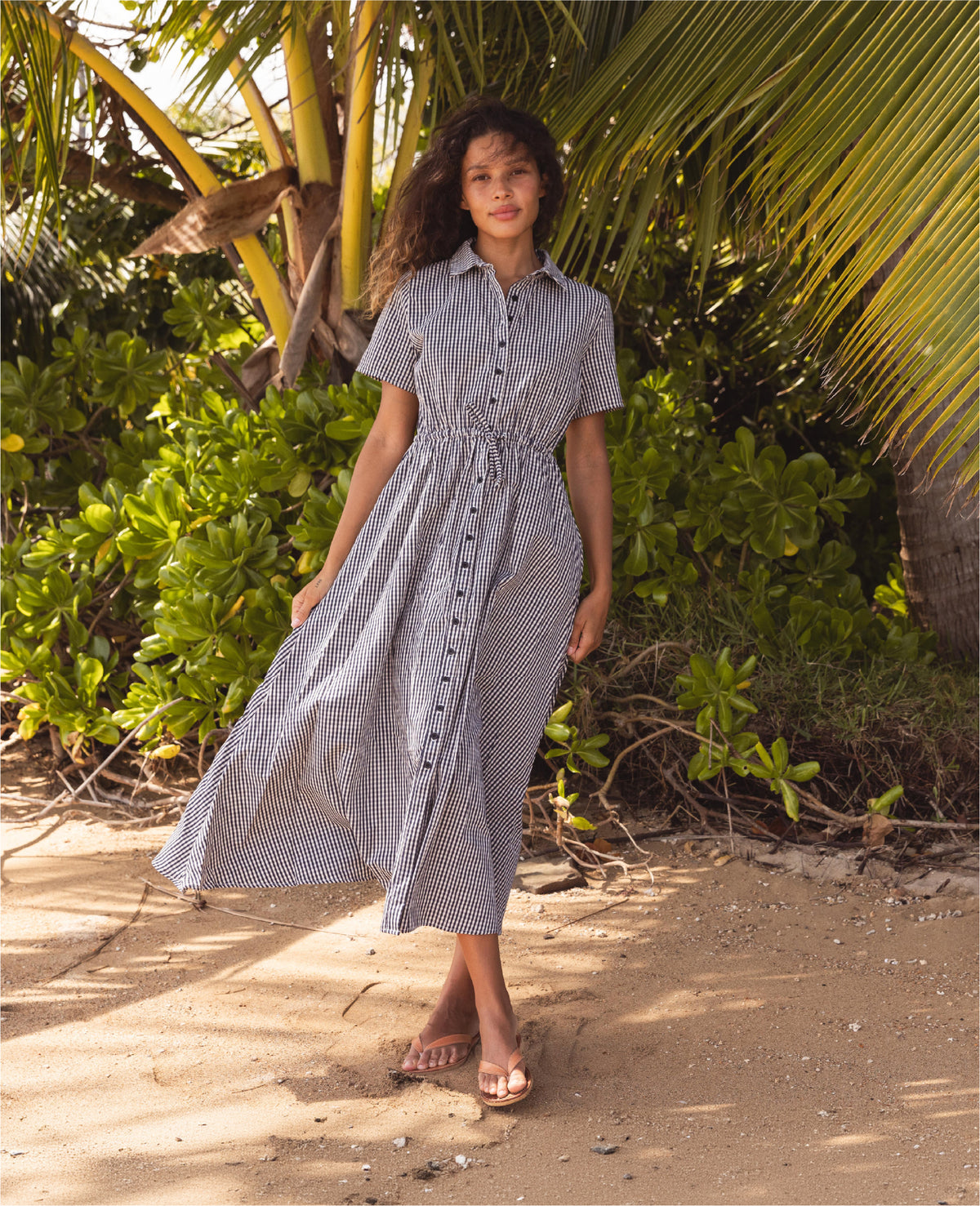 Woman in a blue checkered dress standing in a tropical setting with palm trees.