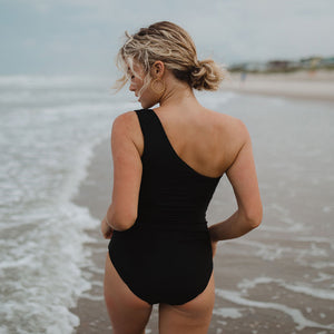 Model in ocean wearing one-shoulder black one-piece swimsuit