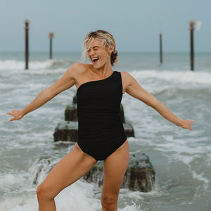 Model having fun in the ocean wearing black one-piece ruched swimwear